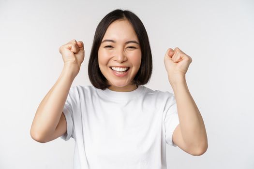 Portrait of enthusiastic asian woman winning, celebrating and triumphing, raising hands up, achieve goal or success, standing over white background.