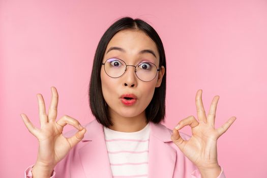 Close up portrait of business woman looks impressed and shows okay sign in approval, recommending company. Young corporate lady in glasses shows ok gesture, pink background.