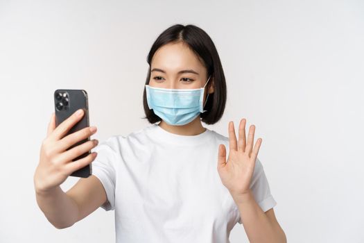 Health and covid-19 concept. Modern asian girl, student in medical mask, video chat with mobile phone, waving hand at smartphone app, standing over white background.