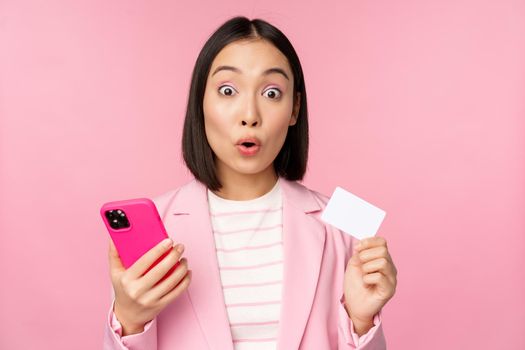 Amazed businesswoman, asian girl in suit showing credit card and mobile phone, order online, shopping with smartphone, standing over pink background. Advertising concept.