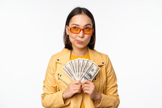 Microcredit and money concept. Stylish asian young woman in sunglasses, laughing happy, holding dollars cash, standing over white background.