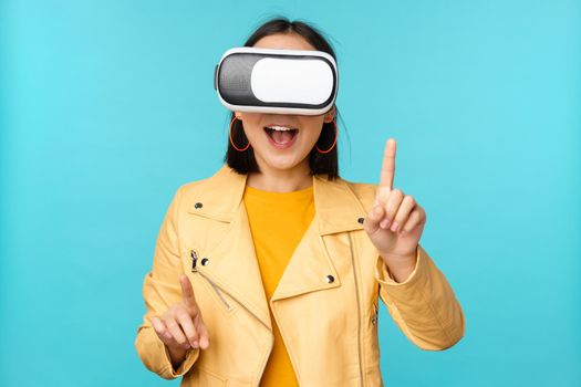 Young asian woman using virtual reality glasses, using VR headset, standing amused against blue background. Copy space