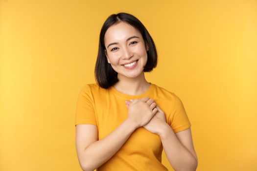 Romantic korean girl, asian woman holding hands on heart, smiling with care and tenderness, standing over yellow background.