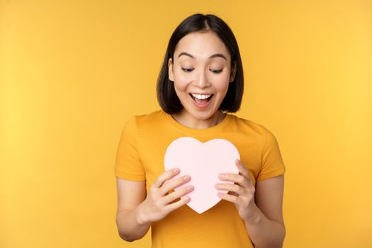 Romance and valentines day. Happy beautiful asian woman holding big heart card and smiling, standing over yellow background.