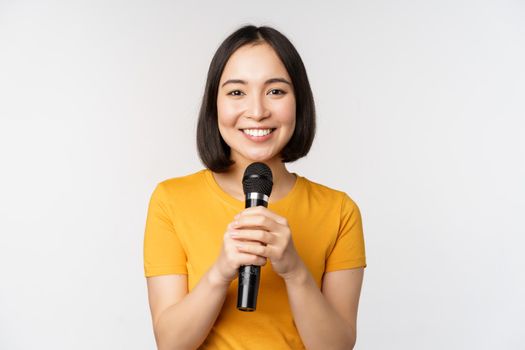 Image of young asian woman talking in microphone, perfom with mic, giving speech, standing in yellow tshirt against white background.