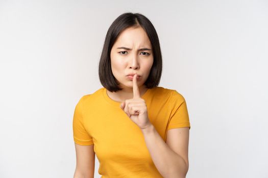 Portrait of angry asian woman shushing, press finger to lips, taboo stop talking sig, looking annoyed, scolding, standing over white background.