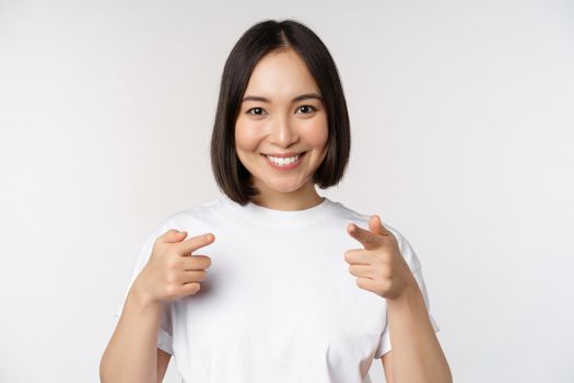 Image of smiling asian girl pointing fingers at camera, choosing, inviting you, congratulating, standing in tshirt over white background.