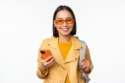 Stylish young asian woman tourist, traveller with backpack and smartphone smiling at camera, posing against white background. Copy space