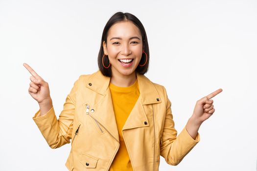 Happy beautiful asian girl pointing fingers left and right, showing banner logo, demonstrating sale, standing in yellow jacket over white background.