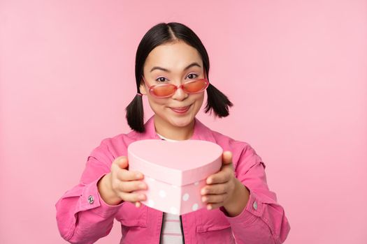 Cute asian girl giving you gift in heart shaped box, kissing and smiling, concept of holiday and celebration, standing over pink background.