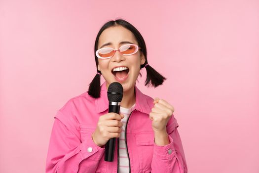 Happy beautiful asian girl singing with mic, using microphone, enjoying karaoke, posing against pink studio background.