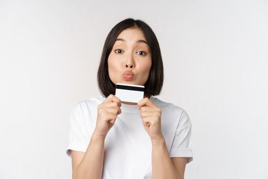 Money and finance concept. Cute japanese girl kissing her credit card, standing in tshirt over white background.