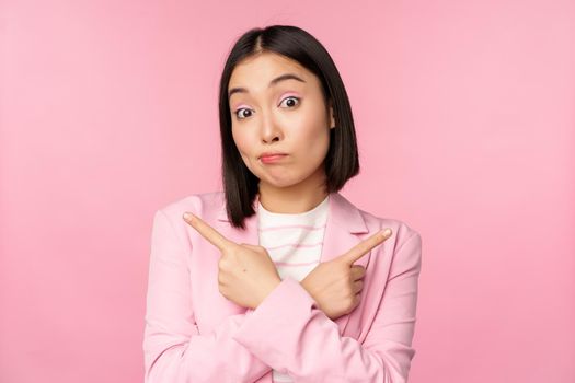 Decision concept. Young asian businesswoman, corporate worker pointing fingers sidways, two directions, showing variants, looking indecisive while choosing, pink background.