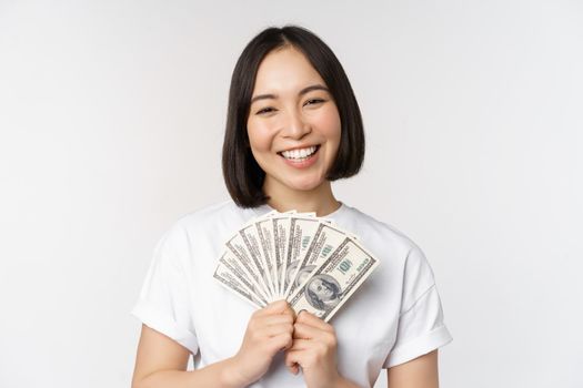 Portrait of smiling asian woman holding dollars money, concept of microcredit, finance and cash, standing over white background.