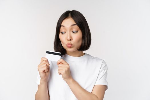 Money and finance concept. Cute japanese girl kissing her credit card, standing in tshirt over white background.