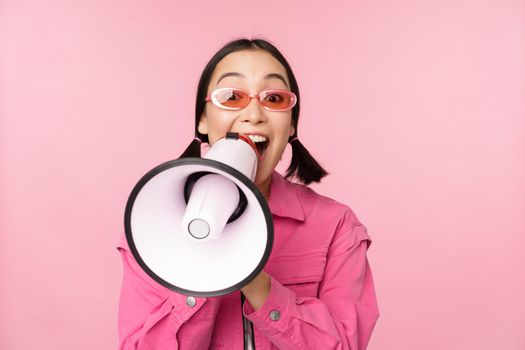 Attention, announcement concept. Enthusiastic asian girl shouting in megaphone, advertising with speaker, recruiting, standing over pink background.