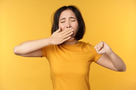 Image of young asian girl student, yawning, feeling tired, standing sleepy agaisnt yellow background. Copy space