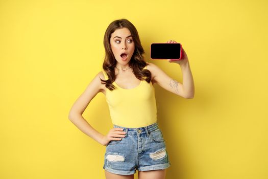 Shocked and amazed stylish girl, showing smartphone screen, app on horizontal mobile phone display, standing in summer clothes over yellow background.