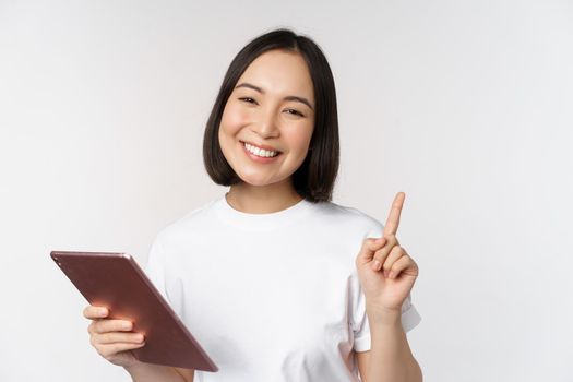 Enthusiastic asian woman with tablet, raising finger and looking amazed, pointing up, standing over white background.