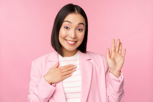 Portrait of cute asian businesswoman raising hand, introduce herself in office, smiling coquettish, standing over pink background in suit.