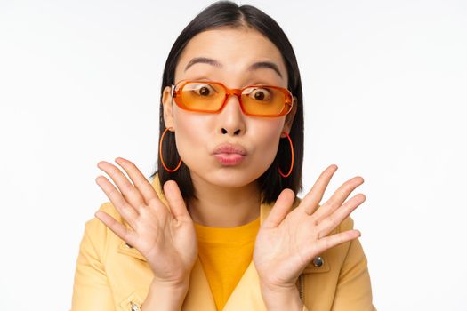 Stylish young asian woman in trendy sunglasses, pucker lips silly, looking surprised, impressed reaction, standing over white background.