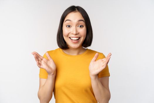 Close up portrait of asian woman looking surprised, wow face, staring impressed at camera, standing over white background in yellow t-shirt.