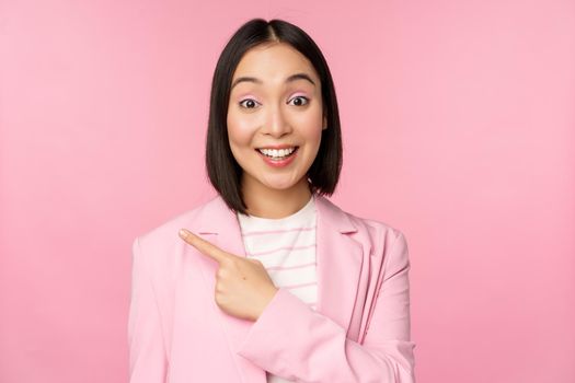 Portrait of asian business woman, saleswoman in suit pointing finger left, showing banner advertisement, smiling and looking professional, pink background.