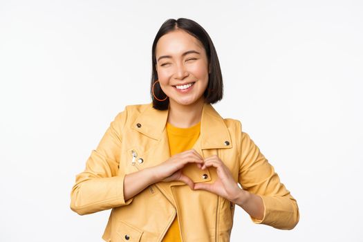 Image of beautiful asian girl showing heart, love sign, express care and romantic feelings, standing over white studio background.