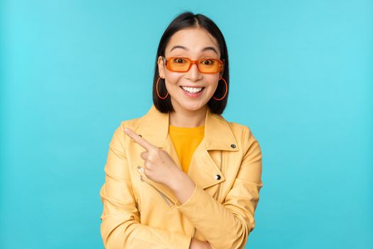 Stylish smiling asian woman in sunglasses, pointing finger left, showing advertisement banner, standing in trendy yellow coat over blue background.