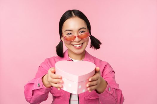 Cute asian girl giving you gift in heart shaped box, kissing and smiling, concept of holiday and celebration, standing over pink background.