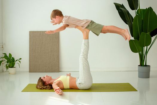 Happy slim woman doing yoga with a little boy lies on a gym mat, in the room, lifting her legs up, doing exercises with support. copy space.