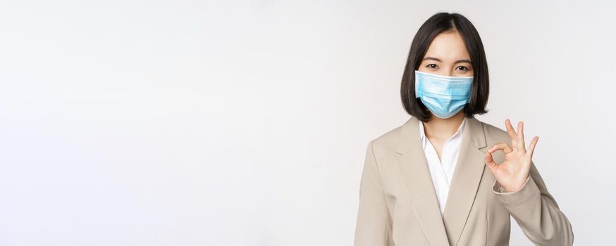 Coronavirus and workplace concept. Image of asian saleswoman, company worker in medical mask showing okay sign, smiling pleased, white background.