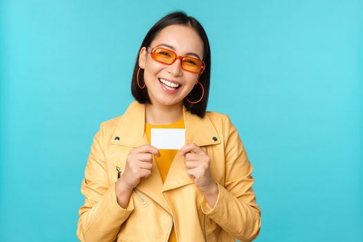 Stylish young asian woman in sunglasses, showing credit card and smiling, recommending bank, contactless payment or discounts in store, standing over blue background.