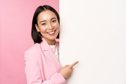 Image of asian female entrepreneur in suit, pointing finger at board, showing smth on white wall, demonstrating chart or information, standing over pink studio background.