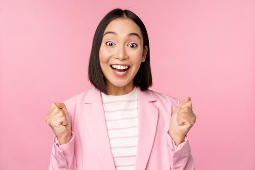 Enthusiastic saleswoman, asian corporate woman say yes, achieve goal and celebrating, triumphing, looking with rejoice and smiling, standing over pink background.