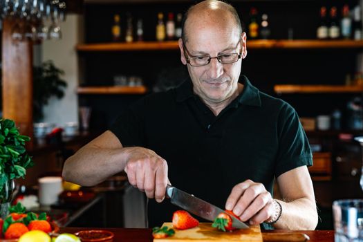 Professional bartender cutting fruit for a mojito. Waiter at the bar preparing drinks. Businessman in his restaurant with traditional ambience and backlighting, bottles of liquor in the background on shelves and fruit on the bar. Bald man with glasses preparing mojito.