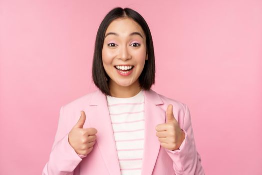 Portrait of asian businesswoman smiling satisfied, showing thumbs up, praise, like and approve, standing in suit over pink background.