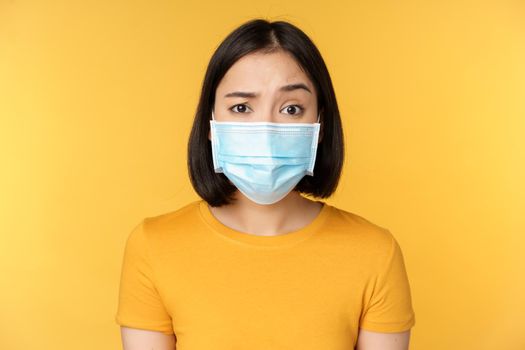 Portrait of skeptical and confused asian woman in medical face mask, raising eyebrow doubtful, standing over yellow background.