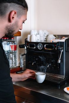 Young waiter with long hair, concentrated and hard-working, dressed in company uniform, a black polo shirt, preparing a coffee in the bar's cafeteria. Restaurant. Warm atmosphere and dim lighting. Steaming cups of coffee.