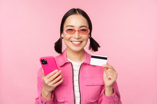 Online shopping. Smiling asian girl shopper, holding smartphone and credit card, paying in mobile app, standing over pink background.