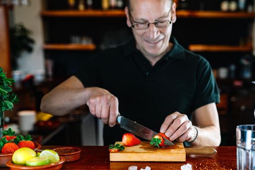 Professional bartender cutting fruit for a mojito. Waiter at the bar preparing drinks. Businessman in his restaurant with traditional ambience and backlighting, bottles of liquor in the background on shelves and fruit on the bar. Bald man with glasses preparing mojito.