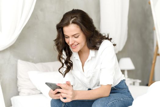 Happy young woman with mobile phone sitting on bed and browsing social media at home