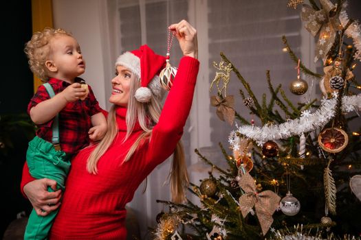 In the living room, a mother holds her son in her arms and a Christmas tree bauble in an expensive one. Boy with curly blonde hair. Santa's hat.