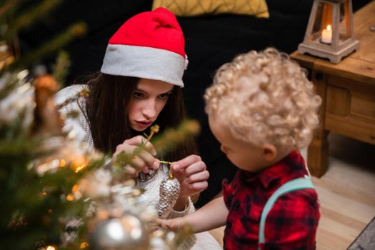 A teenage girl and her brother decorate a Christmas tree for Christmas. A two-year-old child with curly blonde hair.