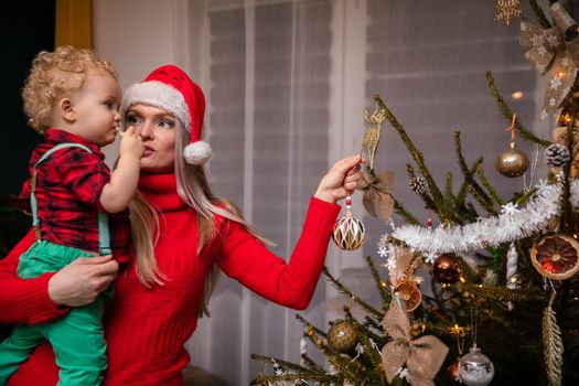 In the living room, a mother holds her son in her arms and a Christmas tree bauble in an expensive one. Boy with curly blonde hair. Santa's hat.