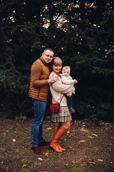 Portrait of attractive young mother and handsome smiling father wearing glasses holding their beautiful lovely baby girl on hands standing against green hedge in autumnal park. They are smiling and looking at camera.