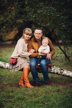 Portrait of attractive young mother and handsome smiling father wearing glasses holding their beautiful lovely baby girl on hands standing against green hedge in autumnal park. They are smiling and looking at camera.