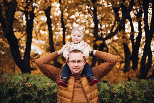 Portrait of strong father wearing glasses, brown jersey and vest holding his adorable daughter while she sitting on his shoulders smiling away with calm face against golden trees in background. They are standing in the autumnal forest.