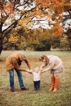 Portrait of attractive young mother and handsome smiling father wearing glasses holding their beautiful lovely baby girl on hands standing against green hedge in autumnal park. They are smiling and looking at camera.