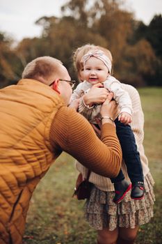 Portrait of attractive young mother and handsome smiling father wearing glasses holding their beautiful lovely baby girl on hands standing against green hedge in autumnal park. They are smiling and looking at camera.